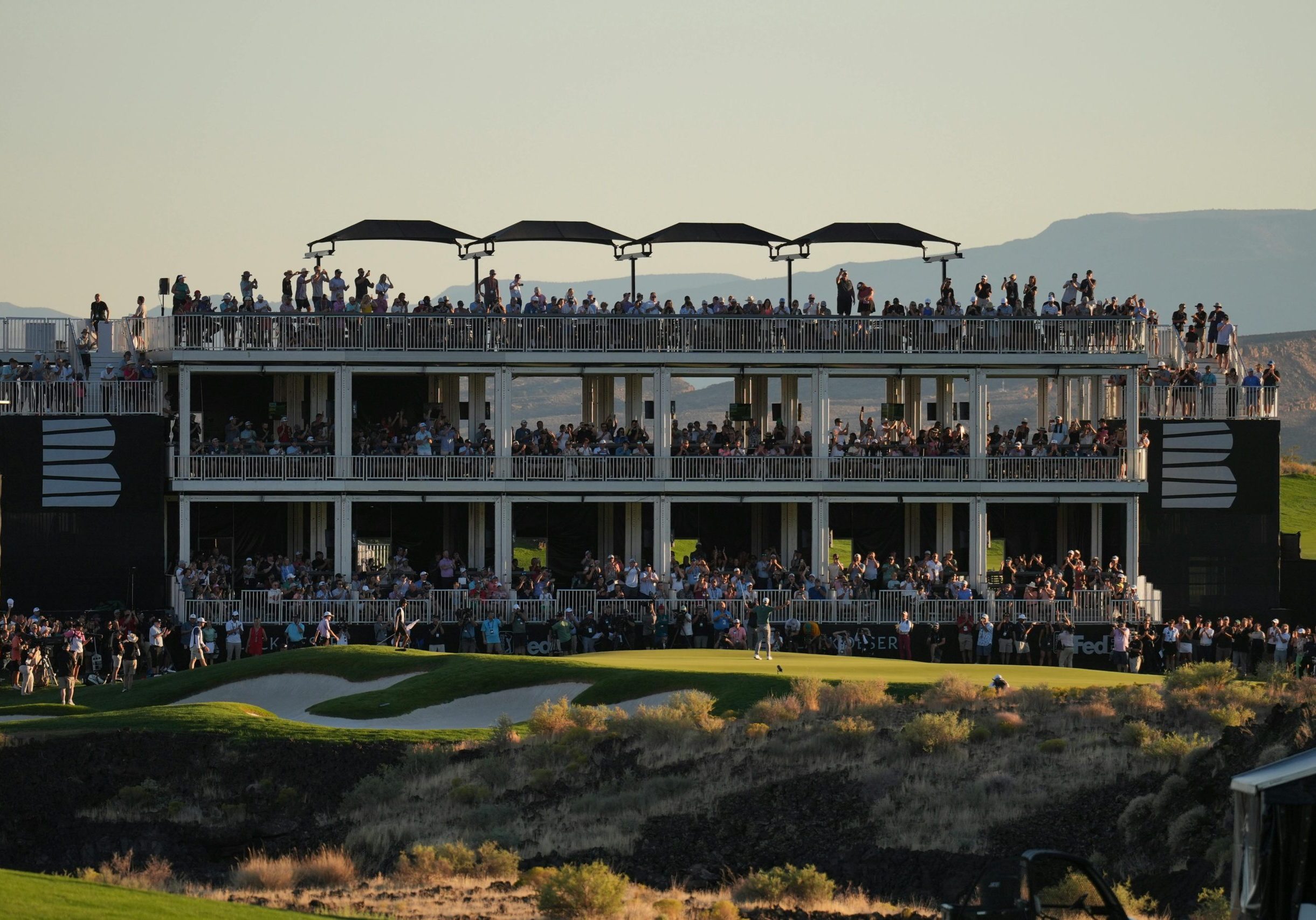 Large crowd gathers on multi-level viewing structure at a golf event during sunset.